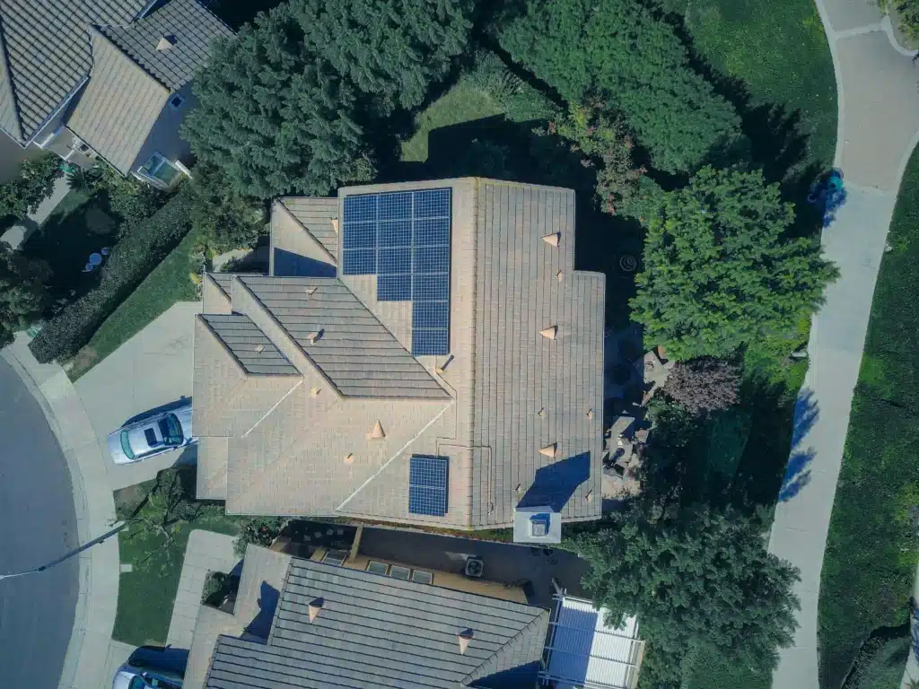 Aerial view of a house with solar panels installed on the roof, surrounded by trees and neighboring homes in a suburban area.