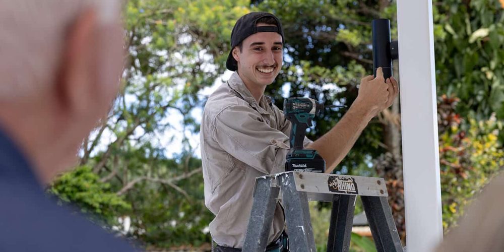 A cheerful Allure Energy electrician installing a security camera outdoors in Cairns, observed by a client.